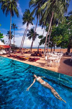 Woman swimming floating in the pool in a tropical seaside resort on the beach. Deck chairs and umbrellas, palm trees and sky. Sea holidays.