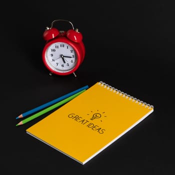 alarm clock with notepad and colored pencils on black background, isolated. back to school. great ideas
