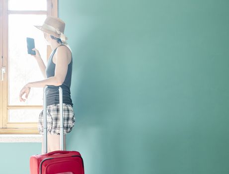 Woman in skirt, with straw hat and striped shirt prepared with a red suitcase and her face mask for her vacation in Covid's time