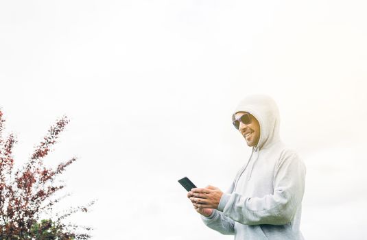 Young man in gray sweatshirt and blue sunglasses holding cell phone in his hands and smiling with trees and a white background