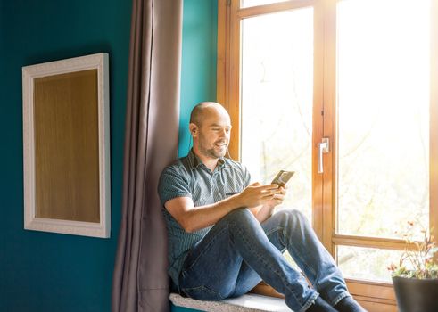 Man sitting at the window holding a mobile phone in his hands and using a wireless headphones