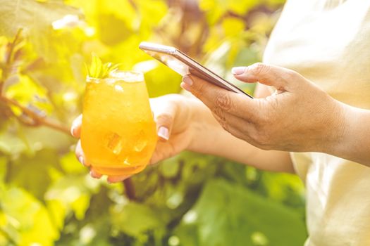 Woman hands using mobile phone and holding refreshing orange drink with ice on summer sunny garden background. Technology in our life at summer time.