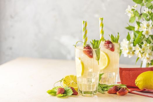 Two glasses of cold icy refreshing drink with lemon and strawberry served with bar tools on light pink table with white blooming flowers. Fresh cocktail drinks with ice fruit and herb decoration.