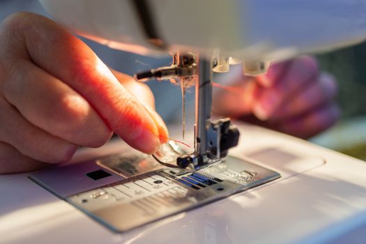 elder woman fingers inserting a red thread into a sewing machine needle, close-up with selective focus