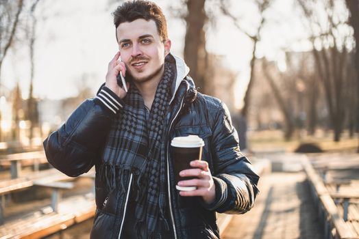 Portrait of attractive man in wearing jacket using mobile phone and holding takeaway coffee while walking through city street.