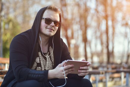 The Man in white headphones and black hood works and communicates on a tablet computer in the park. Closeup of a guy in a black hood and a tablet in his hands running on a tablet.