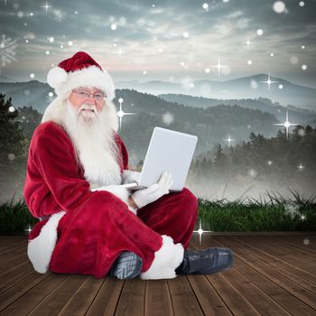 Santa sits and uses a laptop against mountain range beyond wooden floor
