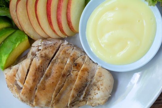 Top view grilled chicken breast steak with sliced avocado, red apple served with salad dressing in white bowl on dish in cafe and restaurant.