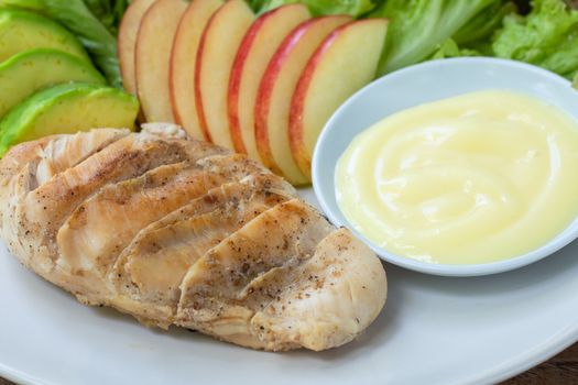 Close up grilled chicken breast steak with sliced avocado, red apple and green lettuce leaves served with salad dressing in white bowl on dish in cafe and restaurant.