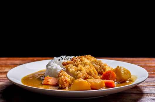 Japanese curry with rice, black sesame, carrot, potato and deep fries chicken in white dish on wooden table.