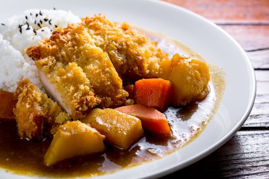 Japanese curry with rice, black sesame, carrot, potato and deep fries chicken in white dish on wooden table.