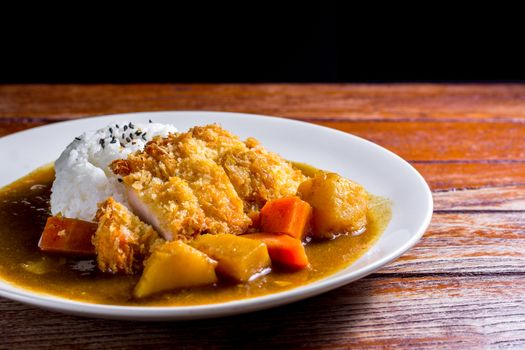 Japanese curry with rice, black sesame, carrot, potato and deep fries chicken in white dish on wooden table.