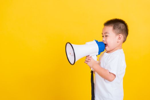 Asian Thai happy portrait cute little cheerful child boy holding and shouting or screaming through the megaphone her looking to side, studio shot isolated on yellow background with copy space