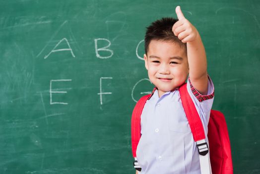 Back to School. Happy Asian funny cute little child boy kindergarten in student uniform with school bag smiling show finger thumb up on green school blackboard, First time to school education concept