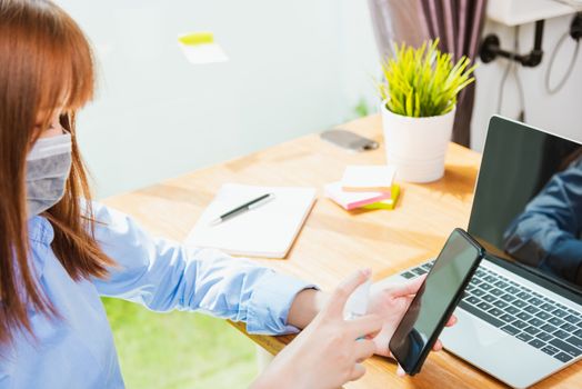 Asian young business woman wearing face mask protective working from home office with laptop computer he quarantines disease coronavirus or COVID-19 using sprays sanitizer cleaning smart mobile phone