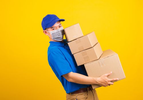 Asian young delivery worker man in blue t-shirt and cap uniform wearing face mask protective lifting stack a lot of boxes, under coronavirus or COVID-19, studio shot isolated yellow background