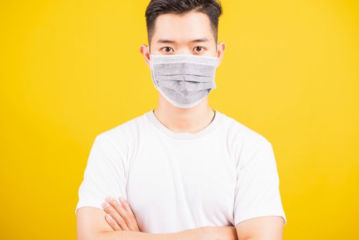 Closeup Asian young handsome man wearing face mask protective germ virus and stand crossed arm, studio shot isolated on yellow background and copy space, medical outbreak coronavirus COVID-19 concept