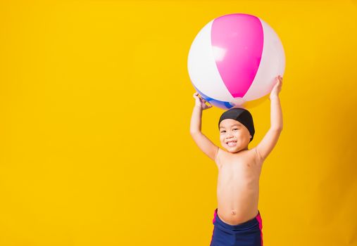 Summer vacation concept, Portrait Asian happy cute little child boy smiling in swimsuit hold beach ball, Kid having fun with inflatable ball in summer vacation, studio shot isolated yellow background