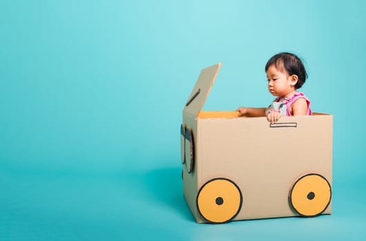 Happy Asian Baby girl smile in driving play car creative by a cardboard box imagination, summer holiday travel concept, studio shot on blue background with copy space for text