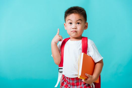 Back to school concept. Portrait Asian cute little child boy face serious hug books thinking and point finger up space, isolated blue background. Kid from preschool kindergarten with school bag