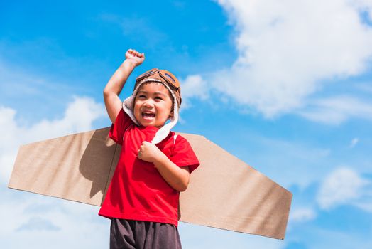 Happy Asian funny child or kid little boy smile wear pilot hat and goggles play toy cardboard airplane wing flying raises hand up against summer blue sky cloud background, Startup freedom concept