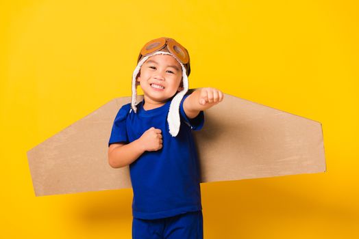 Happy Asian handsome funny child or kid little boy smile wear pilot hat play and goggles raise hand up with toy cardboard airplane wings flying, studio shot isolated yellow background, Startup freedom