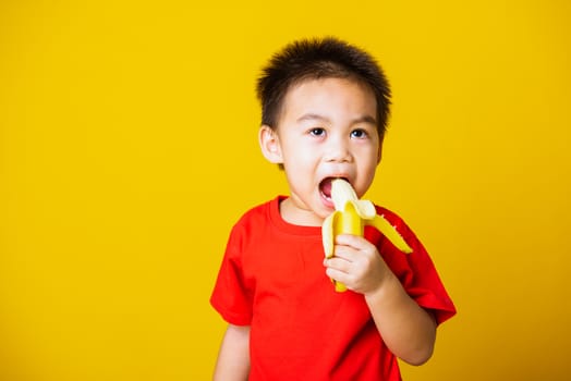 Happy portrait Asian child or kid cute little boy attractive smile wearing red t-shirt playing holds peeled banana for eating, studio shot isolated on yellow background