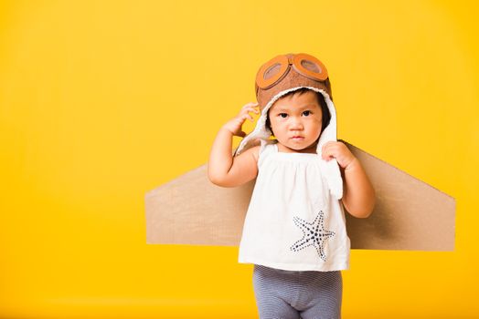 Happy Asian beautiful funny baby little girl smile wear pilot hat raise hand up play and goggles with toy cardboard airplane wings fly, studio shot isolated yellow background, Startup freedom concept