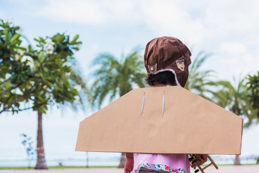 Happy Asian funny child or kid little girl smile wear pilot hat and goggles play toy cardboard airplane wing flying against summer sky cloud on trees garden background, Startup freedom concept