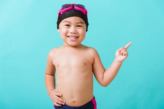 Summer vacation concept, Closeup portrait Asian happy cute little child boy wear goggles and swimsuit, Kid having fun with in summer vacation point to side away, studio shot isolated blue background