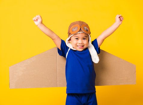 Happy Asian handsome funny child or kid little boy smile wear pilot hat play and goggles raise hand up with toy cardboard airplane wings flying, studio shot isolated yellow background, Startup freedom