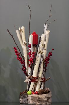 Christmas red candle decorated with a bunch of wooden twigs and berries and fir cones on a gray background