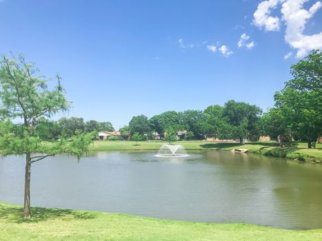 Floating pond fountain at local park in Coppell, Texas, America. Watering decorative fountain in green park with mature trees and residential houses in background.