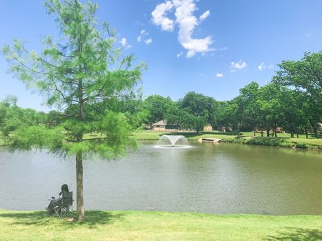 Rear view Asian lady sitting on folding chair fishing at local park with watering fountain in Coppell, Texas, America. Recreational activity active lifestyle at residential park under sunny cloud blue sky.