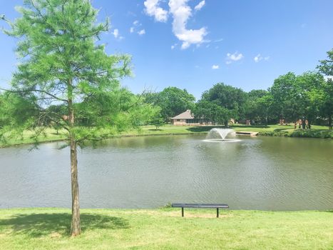 Empty metal bench looking at clear pond with working floating water fountain at residential park in Coppell, Texas, America. Lush green mature trees and single family houses in background