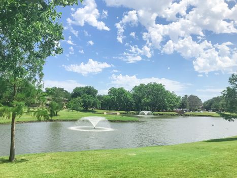 Double floating fountains at residential park in Coppell, Texas, America. Beautiful local park with lush trees and green grass lawn under sunny cloud blue sky.