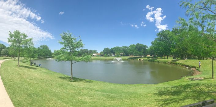 Panorama view residential park with double water fountains on clear pond, pathway and picnic bench under sunny blue cloud sky in Coppell, Texas, USA. Unidentified people fishing and feeding ducks