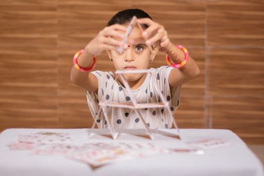 Young girl kid busy in building Pyramid with house of cards at home - concept of focus or concentration excerise game for school children.