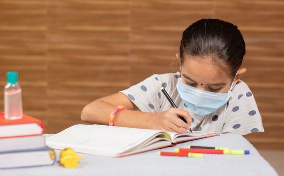 Concept of homeschooling, young girl with medical mask busy in writing at home during covid-19 or coronavirus pandemic lock down