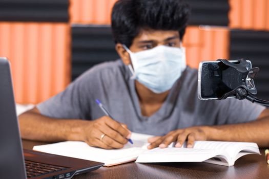 Young man with medical mask e-learning at home due to covid-19 or coronavirus isolation concept - college student taking notes by looking into virtual class on mobile due to quarantine