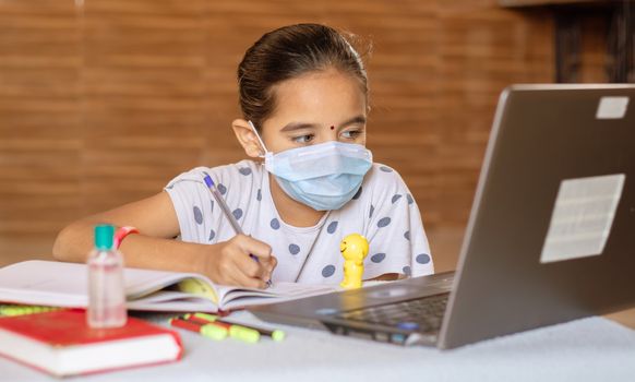 Concept of homeschooling and e-learning, young girl with medical mask writing by looking into laptop during covid-19 or coronavirus pandemic lock down