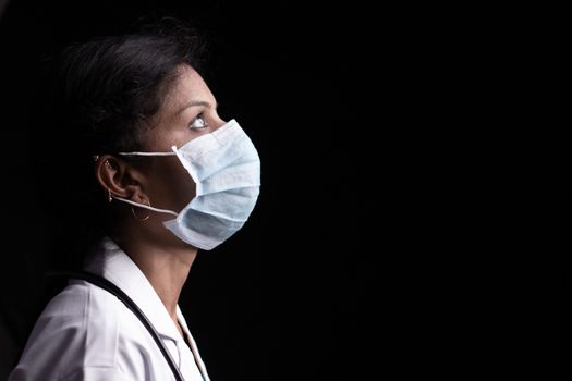 Profile view of young woman doctor with opened eyes in medical mask on black background looking up - concept of hope and fight to end coronavirus or covid-19 crisis