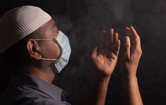 Muslim Man with medical mask and cap praying to god in dark room to protect or save from covid-19 or coronavirus crisis - spirituality and Surrender concept