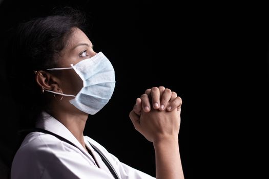 Profile view of young woman doctor in medical mask praying to god on black background looking up - concept of hope and fight to end coronavirus or covid-19 crisis