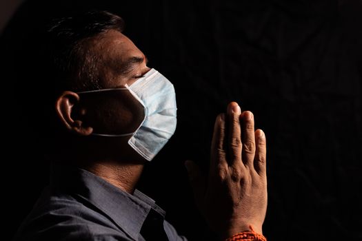 Man with medical mask praying to god by closing eyes in dark room to protect or save from covid-19 or coronavirus crisis - spirituality and Surrender concept.