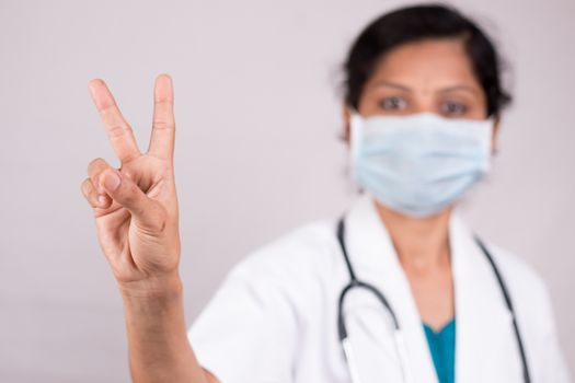 Woman doctor with medical mask in uniform showing V gesture on isolated background - Concept of victory salute or peace gesture by doctor during covid-19 or coronavirus pandemic