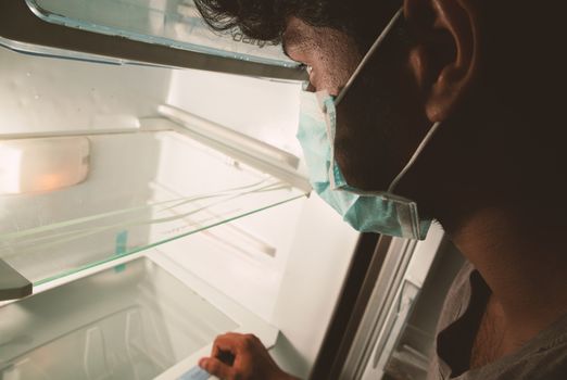 Concept of no pantry food available during home quarantine at covid-19 or coronavirus pandemic - Man in medical mask looking into empty fridge or refrigerator for food