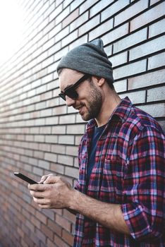 Hipster sms texting phone app in city street on brick wall background. Amazing man holding smartphone in smart casual wear standing. Urban young professional lifestyle.