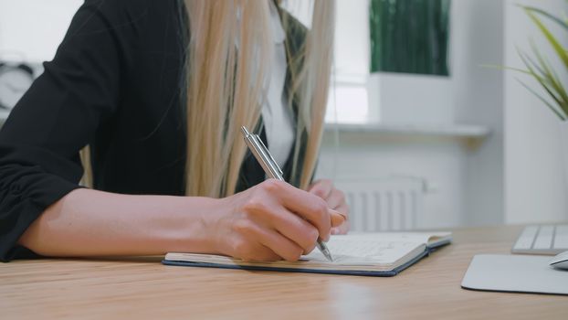 Crop view of woman with long blond hair in bright casual shirt, sitting at wooden desk and writing down information with shiny metal pen into daily planner at day.