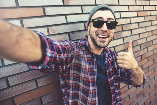 Portrait of stylish handsome young man with bristle standing outdoors and leaning on brick wall. Man wearing sunglasses and hat. Man making selfie.